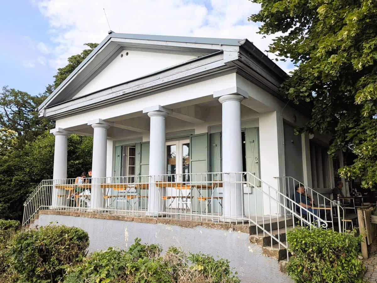 Das historische Kirchberghäuschen, ein weißes Gebäude im klassizistischen Stil mit Säulenportikus und grünen Fensterläden, thronend auf einem Hügel, davor eine Terrasse mit Metallgeländer und Gästen an Tischen.